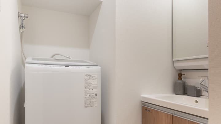 Laundry alcove with a top-loading washing machine next to the bathroom vanity, featuring a sink, mirror, and a wood-grain cabinet base.
