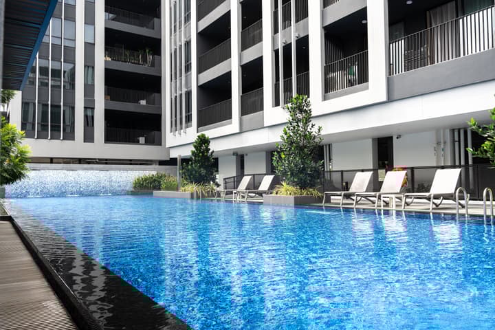 An outdoor swimming pool with vibrant blue water and a small waterfall feature, surrounded by lounge chairs and the modern, black-and-white facade of an apartment building.