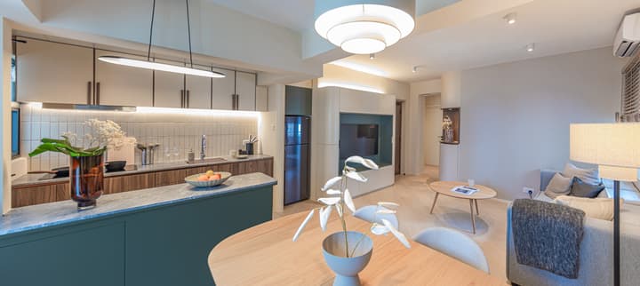 Wide-view of a modern, open-plan apartment featuring a kitchen with a dark green island, a light wood dining table, and a neutral-toned living area with a sofa.