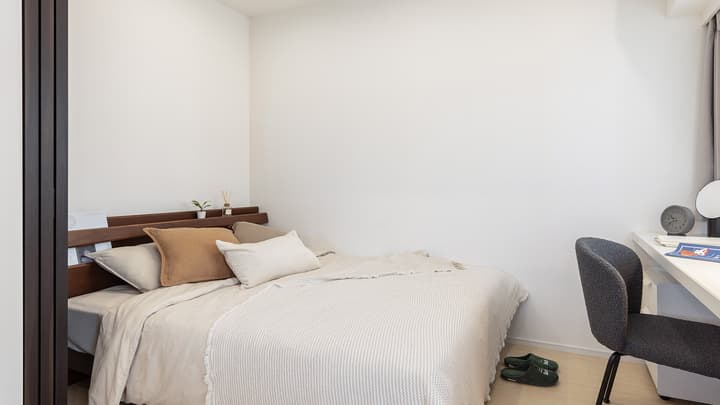 Furnished compact bedroom with a double bed, dark wood headboard, a white desk, and a dark grey chair against a clean white wall.