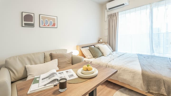 Modern 1K studio apartment featuring a double bed, light beige sofa, small wooden dining table, and abstract wall art, with bright natural light from the window.