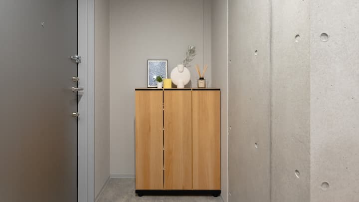 Entrance area of the apartment unit showing a wooden shoe cabinet with decor, a gray entry door, and an adjacent exposed concrete wall.