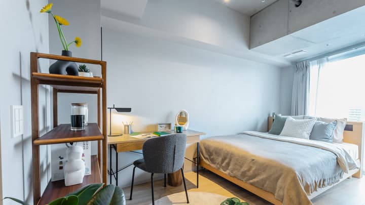 Studio apartment view featuring a light wood bed and a dedicated workspace with a desk, task lamp, and a tall wooden shelf, against a wall with an exposed concrete ceiling element.