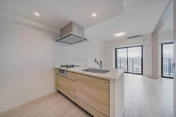 Modern kitchen with light wood cabinets, a white countertop, a stainless steel sink, a cooktop, and a silver range hood, looking toward the open living area and balcony.