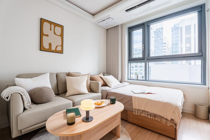 Modern, compact studio apartment with a neutral-toned sofa, a small wooden coffee table, a single bed positioned by a large window, and a bright city view.
