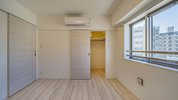 Bright, empty bedroom featuring light wood-style flooring, white walls, an air conditioner, and a light wood sliding door leading to a closet area in a Kunitachi apartment unit.