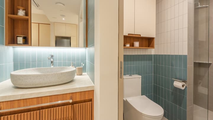 View of the bathroom area with a stone oval basin, fluted wood vanity, and the toilet area with blue and white subway tiles and wooden shelving.