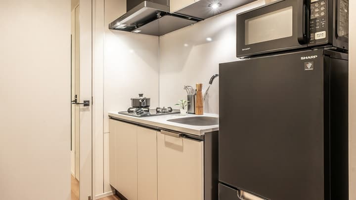Compact galley kitchen featuring a black refrigerator and microwave, light beige cabinets, a sink, and a two-burner cooktop with a pot and pan.