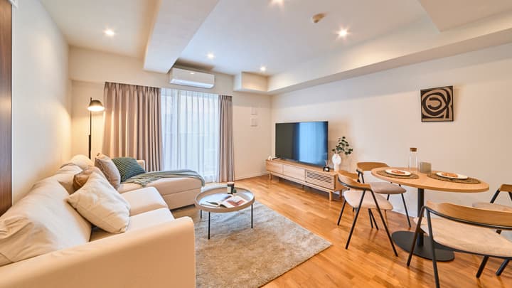 Open-plan living area with a neutral sectional sofa, round coffee table, dining set, media console, and large window with curtains.