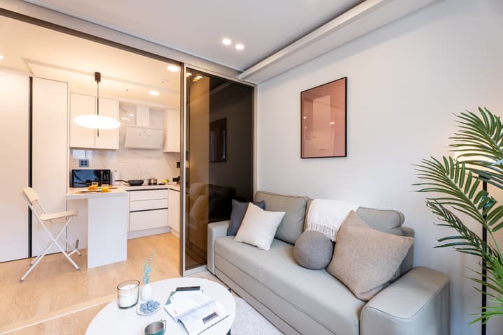 Living room area with a gray sofa and a round coffee table, looking into the adjacent white kitchen/dining area which is separated by a dark-framed sliding glass partition.