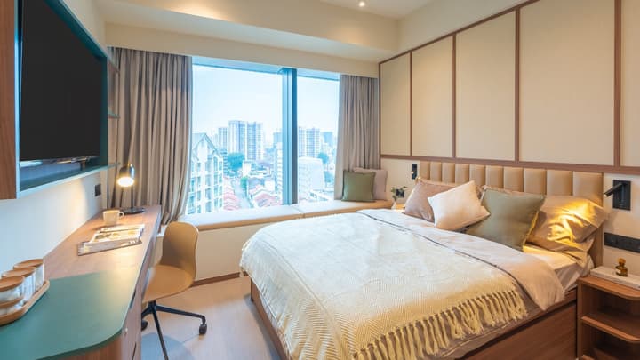 Traditional-style bedroom with a queen bed, upholstered headboard, a large window seat offering a city view, and a small work desk by the window.