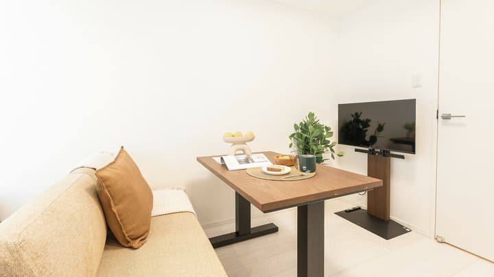 Apartment dining area with a neutral-colored sofa, a dark wood dining table set for one, and a flat-screen television mounted on a sleek, dark wood floor stand.