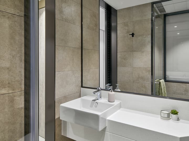 Close-up of a modern bathroom vanity featuring a square white sink, white countertop, a large mirror, and warm beige square wall tiles with black trim.