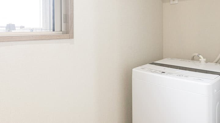 Utility area featuring a white top-loading washing machine beneath a small window, with light beige textured wallpaper on the walls in Monzennakacho.