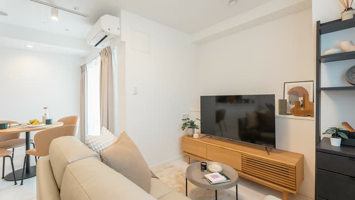View of a modern living area showing a large TV on a slatted wood media console, a light beige sofa, and a black ladder shelf against white walls.