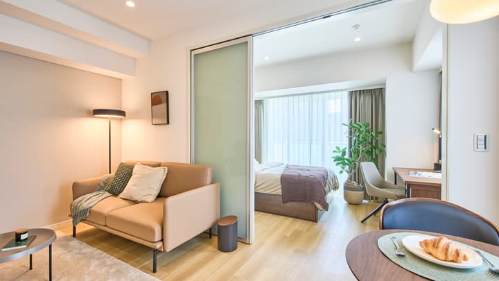 Cozy living area with a leather sofa and floor lamp, looking into the bright bedroom through a frosted glass sliding door.