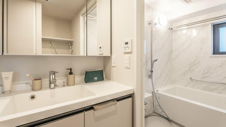 Modern apartment bathroom with a white sink and vanity, a mirrored cabinet, and a shower area with marble-look wall tiling and a small tub.