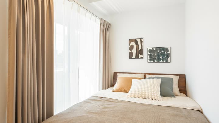 Bedroom area featuring a double bed with a wooden headboard, accent pillows, two framed pieces of abstract wall art, and light-blocking curtains over a large window leading to a balcony.