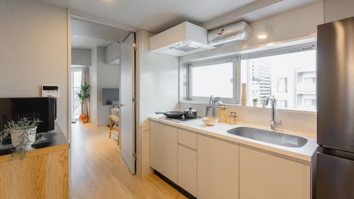 Galley kitchen with white cabinets, a sink, and a cooktop, with a wide window providing a city view. The image shows an open doorway leading to the main living area with wood flooring.