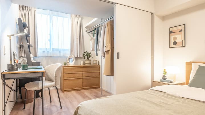 Bright bedroom corner featuring a work desk and chair next to a large window, a light wood dresser, and an open closet area with hanging clothes storage.