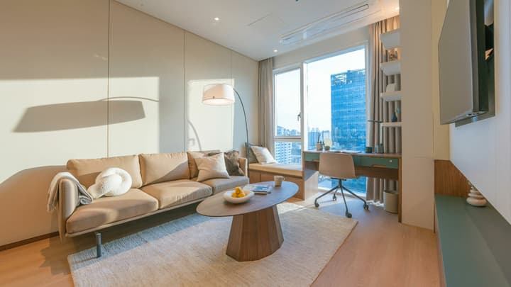 View of the spacious, separate living room in the one-bedroom suite with a modern tan sofa, arc floor lamp, oval coffee table, and a dedicated workspace next to large windows overlooking the city.