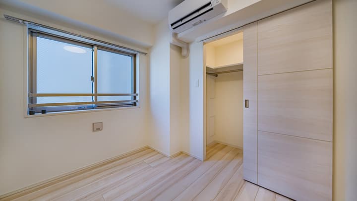 Empty bedroom featuring light wood-style flooring, a window, a wall-mounted air conditioner, and a light wood sliding door opening to a walk-in closet area.