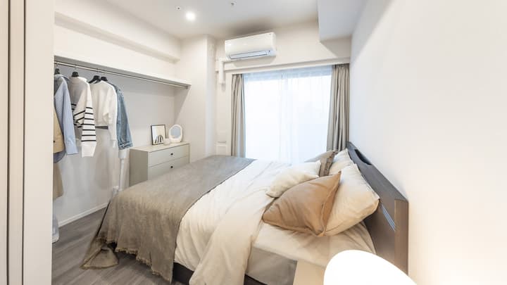 Room Type GalleryaNeutral-toned bedroom with a dark wood bed, light duvet, throw pillows, and an open closet area with hanging clothes and a small dresser below.