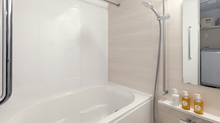 Bright, modern unit bathroom with a deep soaking bathtub, shower, and liquid soap dispensers on a shelf.