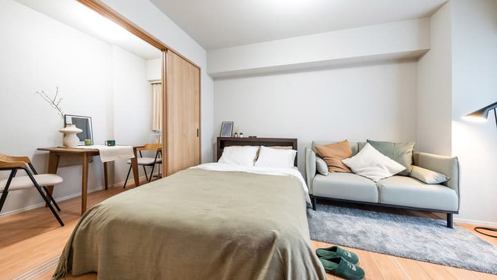 Studio apartment view showing a double bed, small gray sofa, and a partitioned dining nook with a wooden table, featuring light wood-style flooring throughout in Monzennakacho.