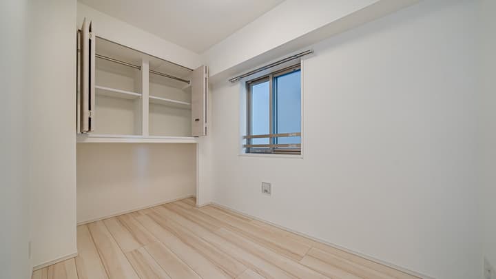 Small, empty room with light wood-style flooring, a bright window, and an open, built-in closet unit with shelving and a hanging rod above a storage niche.