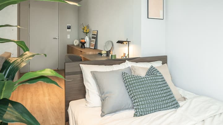 Bedroom view with a wooden bed frame, assorted pillows, and a glimpse of the workspace area with a desk, lamp, and wooden cabinetry separated by the headboard.
