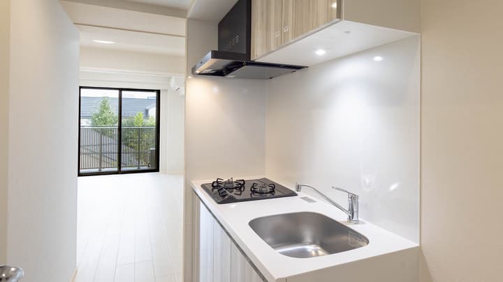 Compact apartment kitchen area with a two-burner black cooktop, stainless steel sink, wood-look cabinetry, and a view into the main living space and balcony.