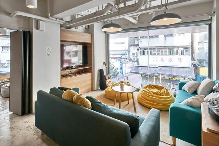 A relaxed, communal lounge space with teal couches and bright yellow beanbag chairs facing a large floor-to-ceiling window overlooking a bustling city street.