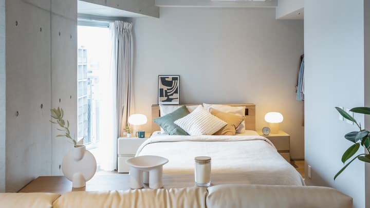 View from the living area over a sofa's backrest toward the bed, showcasing the neutral-toned bedroom with a large window and twin bedside tables with modern lamps.