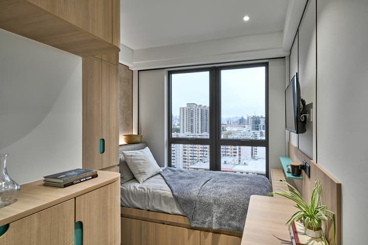 Compact, modern studio room featuring a single bed built into a wooden storage platform, a small integrated desk on the right, a wall-mounted TV, and large windows offering a city view.
