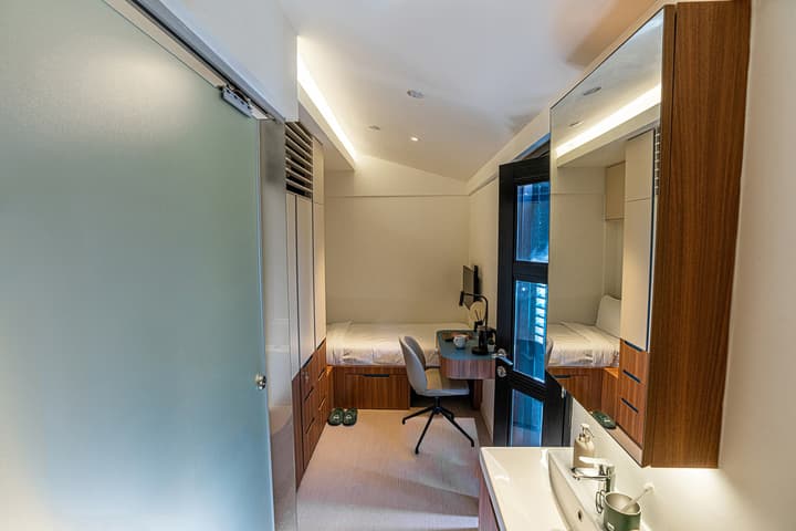 View inside a compact micro-apartment unit featuring a single bed with under-bed storage, a built-in desk, and a small integrated bathroom sink area with a mirrored cabinet.