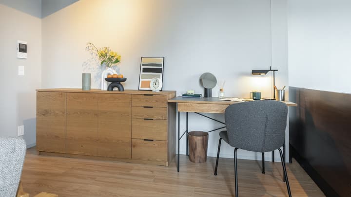 Detailed view of the workspace area featuring a long wooden sideboard for storage, a desk with a dark gray chair, and a modern black task lamp.