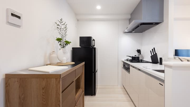 Narrow, minimalist galley kitchen featuring white cabinets, a black fridge, microwave, gas cooktop, and a light wood sideboard with decor.