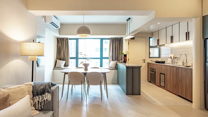 View of the apartment's open-concept dining area with a modern light fixture, window seat, and an adjacent kitchenette with wood and green cabinetry.