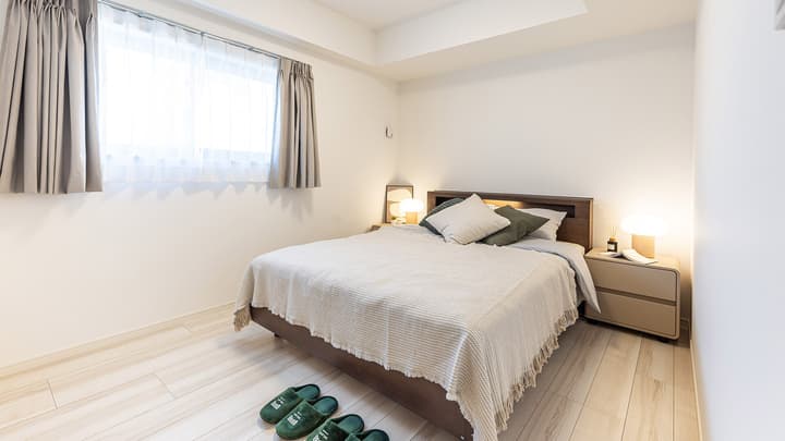 Spacious bedroom with a double bed, light wood frame, matching modern nightstands with glowing lamps, and a large bright window with gray curtains.
