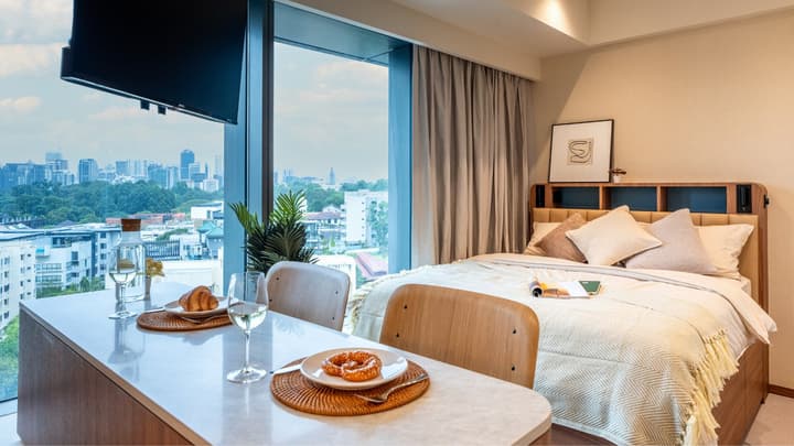 Studio suite with a queen bed, dining bar, and two stools set against a floor-to-ceiling window offering a panoramic city view.