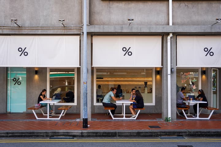 Exterior street view of a cafe with the percentage (%) logo, showing patrons seated at covered outdoor picnic tables along the sidewalk.