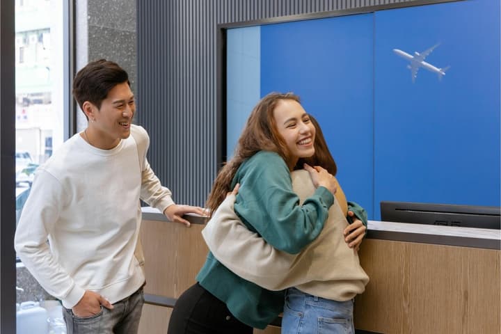 Two smiling women embracing warmly at a bright reception desk while a man looks on. A screen behind them shows an airplane flying.