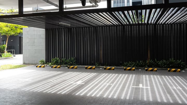 Covered residential car park with black slatted walls and ceiling, creating distinct striped shadows on the paved floor.