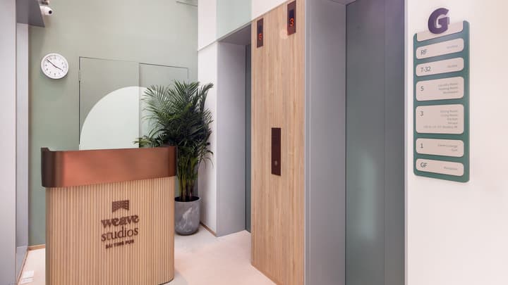 Modern residential lobby with a curved reception desk, wood-paneled elevator, potted plant, and a directory board listing floor amenities like the Gym, Lounge, and Rooftop.