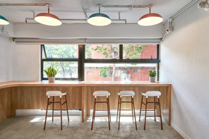 A bright co-working area with a long wooden window counter, featuring four white and wood bar stools and overlooking green trees and a red brick building exterior.