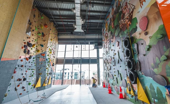 Interior view of a high-ceiling indoor rock climbing gym with walls covered in colorful climbing holds and a cartoon adventure mural on one side.