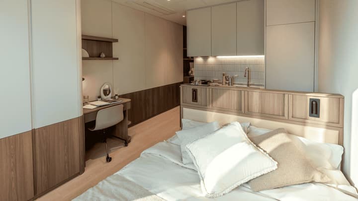 An overall interior shot of the premium suite, showing the desk and wardrobe area in the foreground and the kitchenette with white subway tile backsplash and the double bed separated by a low divider.