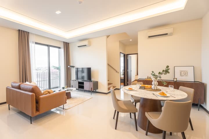 A bright, open-plan living and dining area in a Singapore apartment, featuring a brown leather sofa, a marble dining table, white tiled floors, and a balcony door.