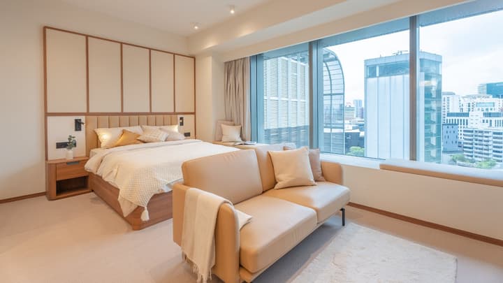 Wide view of a studio apartment showcasing the queen bed with an upholstered headboard, a tan leather loveseat, and full-height city-view windows.